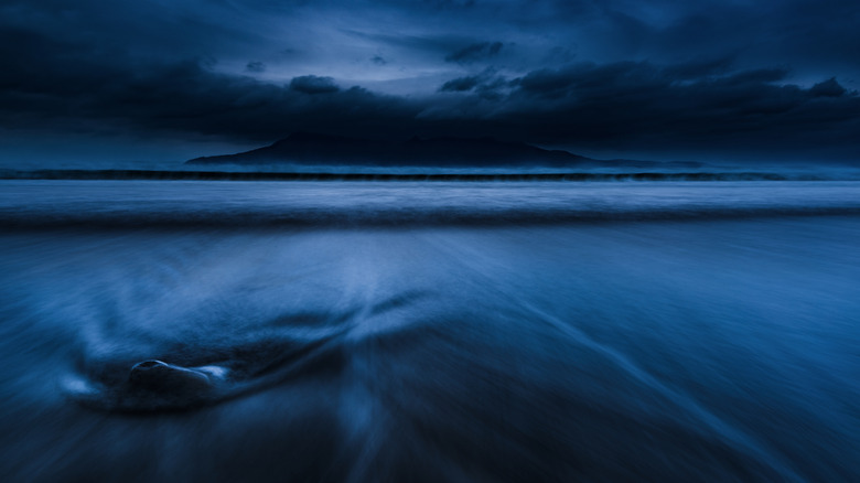 A moody, twilight-blue sky full of clouds, above a beach covered the shifting tide, with an island in the background.