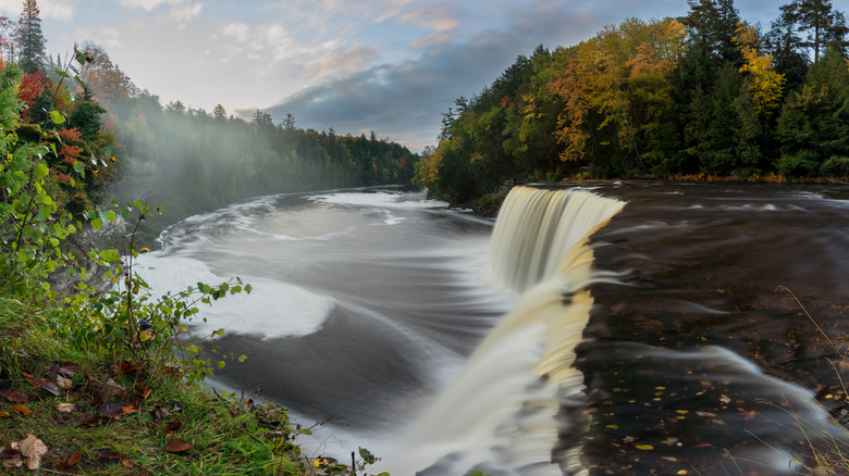 a wide waterfall flows into a river surrounded by fall foliage