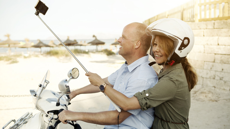 A man and woman ride a scooter with a selfie stick