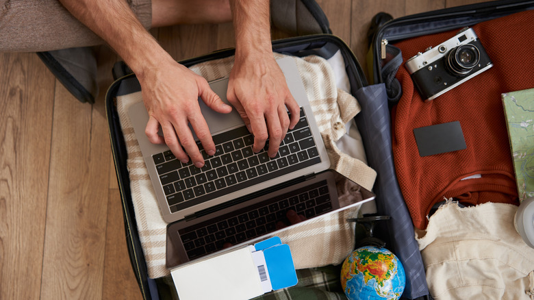 Hands typing on a laptop on top of a packed suitcase.