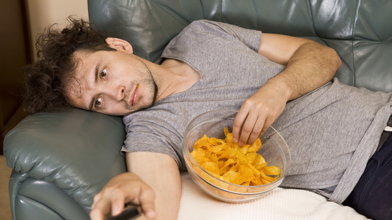 A tired man on a couch eating potato chips.