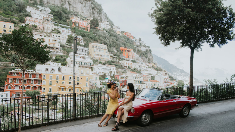 Two women sitting on red car in front of coastal buildings