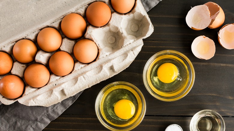 Two eggs cracked in bowls next to a carton of eggs