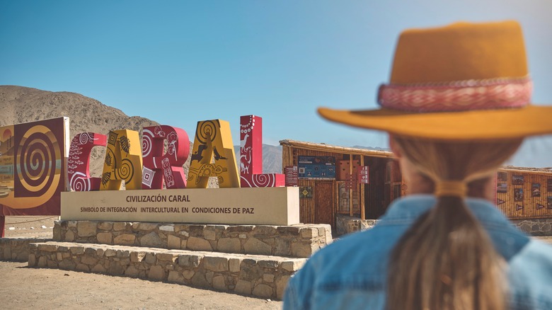 Woman with hat standing in front of Caral sign, Peru