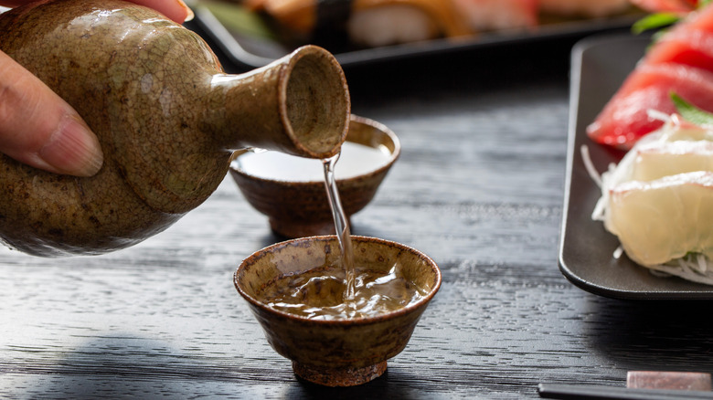 Someone pours sake into a small cup next to plate of sushi