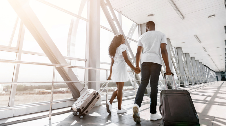 A couple holds hands as they walk through an airport with wheeled luggage.