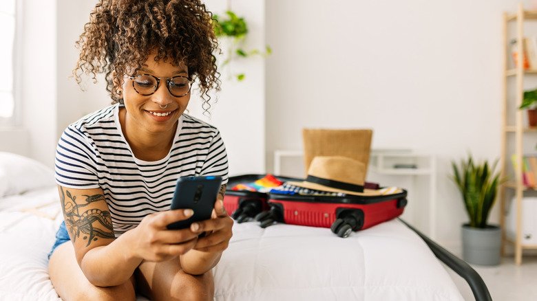 Traveler looking at phone sitting by packed suitcase