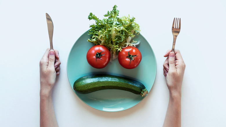 hands holding fork and knife on either side of a plate full of vegetables arranged to look like a face