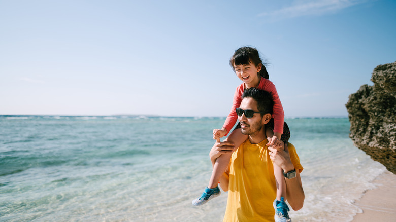 A man carries his daughter on his shoulders on a sunny beach.