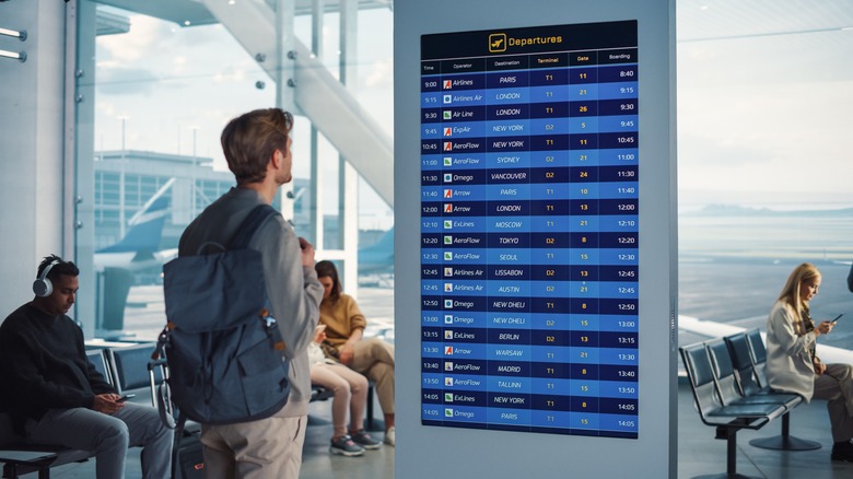 Young Man Looking at Arrival and Departure Information Display Looking for His Flight. Backgrond: Diverse Crowd of People Wait for their Flights in Boarding Lounge of Airline Hub